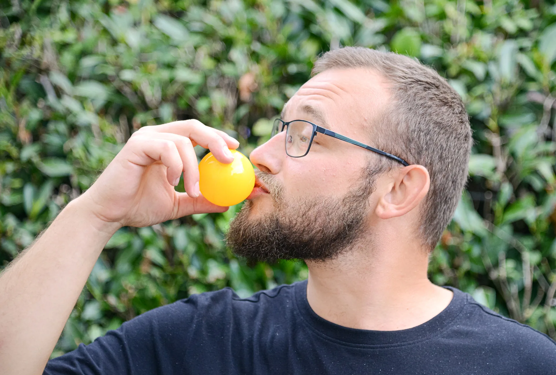 Ein bärtiger Mann mit Brille bläst einen gelben Luftballon vor grünem Blätterhintergrund auf.