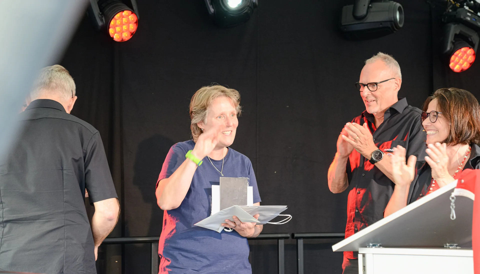 Eine Frau erhält auf einer Bühne eine Urkunde, während zwei Personen applaudieren.