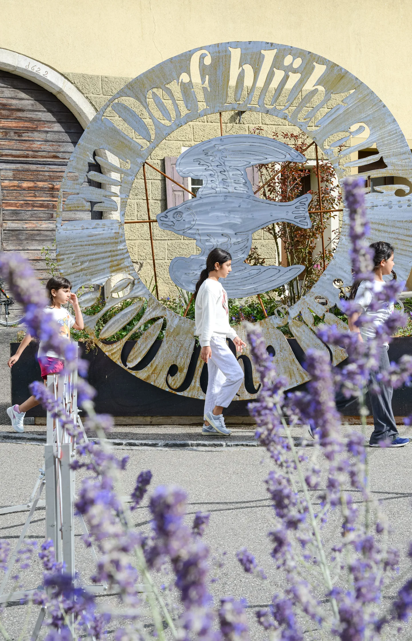 Drei Personen gehen an einem runden, kunstvoll gestalteten Metallschild vorbei, im Vordergrund Lavendelblüten.