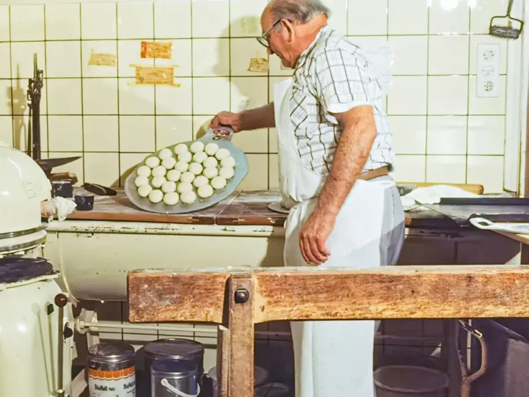 Ein älterer Bäcker mit Brille legt Teiglinge auf ein Blech in einer alten Backstube.