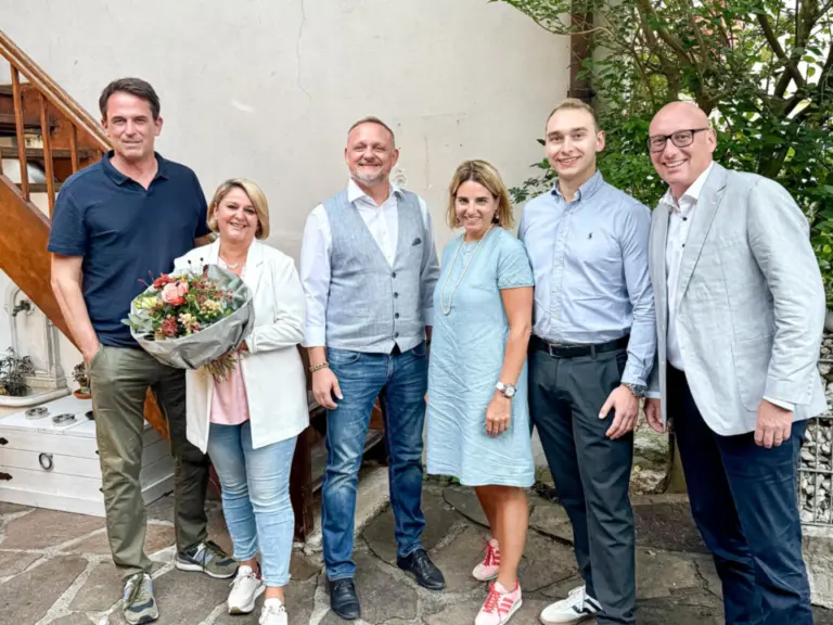 Sechs Personen stehen lächelnd nebeneinander auf einem gepflasterten Hof vor einem Gebäude, eine Frau hält einen Blumenstrauss.