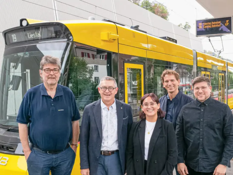 Fünf Personen stehen lächelnd nebeneinander vor einer gelben Tramgarnitur an einer Haltestelle.