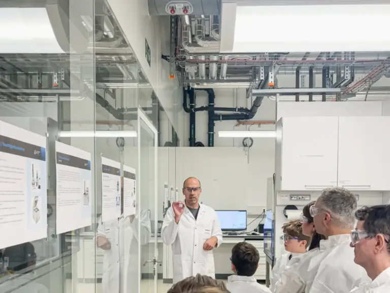 Ein Mann im weissen Labormantel spricht vor einer Gruppe zuhörender Personen in Laborkitteln in einem Labor.