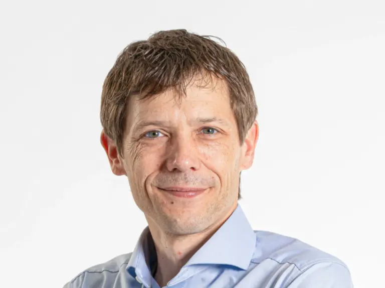 Ein Mann mit verschränkten Armen in hellblauem Hemd und Jeans lächelt vor weissem Hintergrund.