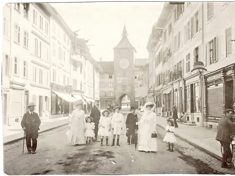Frauen, Männer und Kinder in eleganter alter Kleidung stehen und gehen auf einer Kopfsteinpflasterstrasse zwischen Häuserzeilen mit einem Torturm.