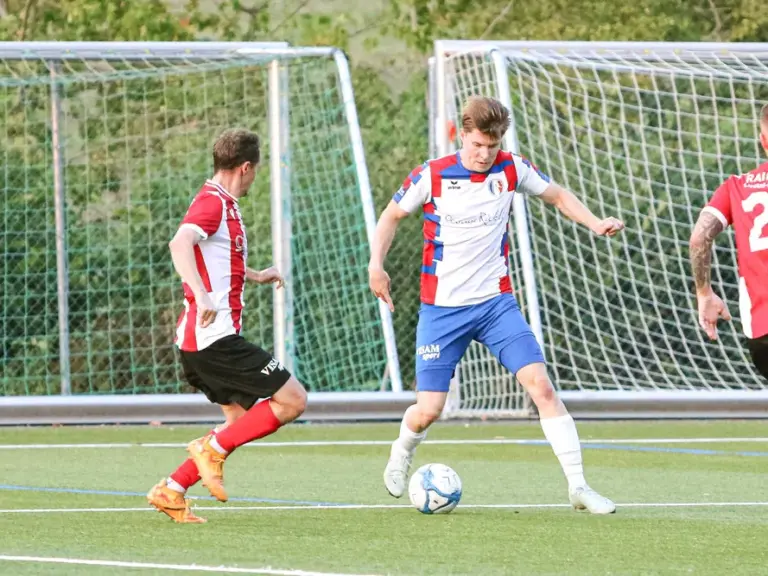 Fussballspieler in blau-roten und rot-weissen Trikots laufen auf dem Rasen einem Ball nach.