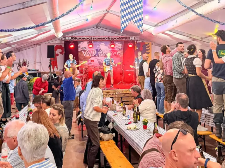 Menschen in Trachten stehen und tanzen auf Bänken in einem festlich geschmückten Festzelt vor einer Bühne.