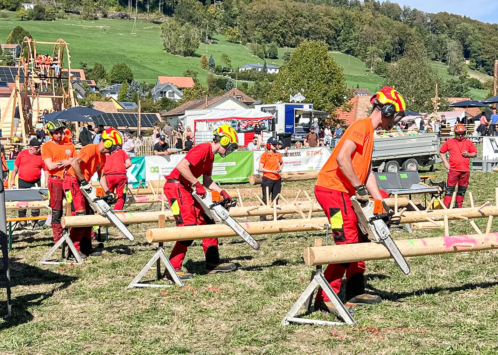 Mehrere Personen in orange-roter Schutzkleidung und Helmen sägen mit Kettensägen Holzstämme auf Böcken.