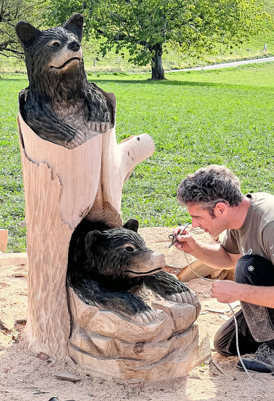 Ein Mann kniet und bearbeitet mit einem Werkzeug eine Holzskulptur aus zwei geschnitzten Bären auf einer Wiese.