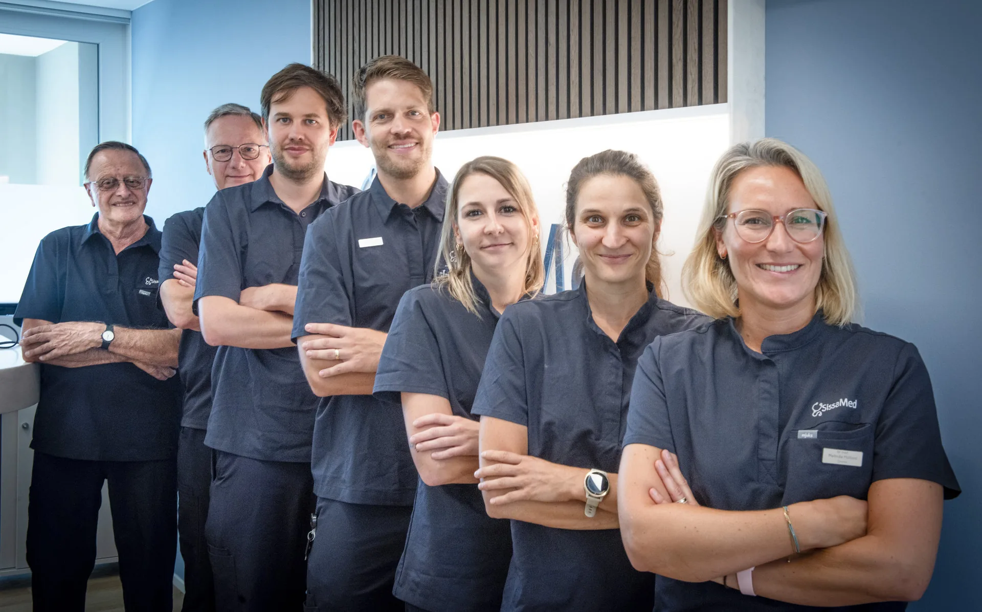 Sieben Personen in dunklen Praxiskitteln stehen aufgereiht und lächeln in die Kamera, viele mit verschränkten Armen.