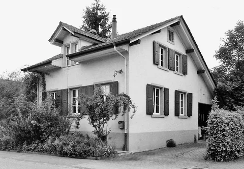 Schwarz-weisses Foto eines zweistöckigen Hauses mit grünen Fensterläden, umgeben von Sträuchern und Bäumen.