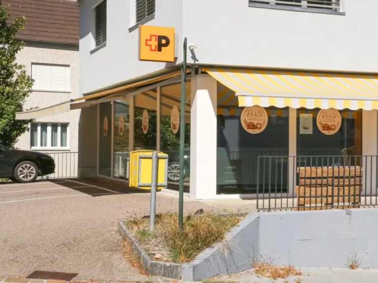 Gelb-weisses Ladengeschäft mit Markise und Post-Schild an einem Wohnhaus.