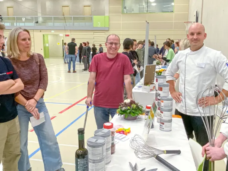 Zwei Köche in weisser Kleidung stehen an einem Tisch mit Kochutensilien und sprechen mit Besuchern in einer Halle.