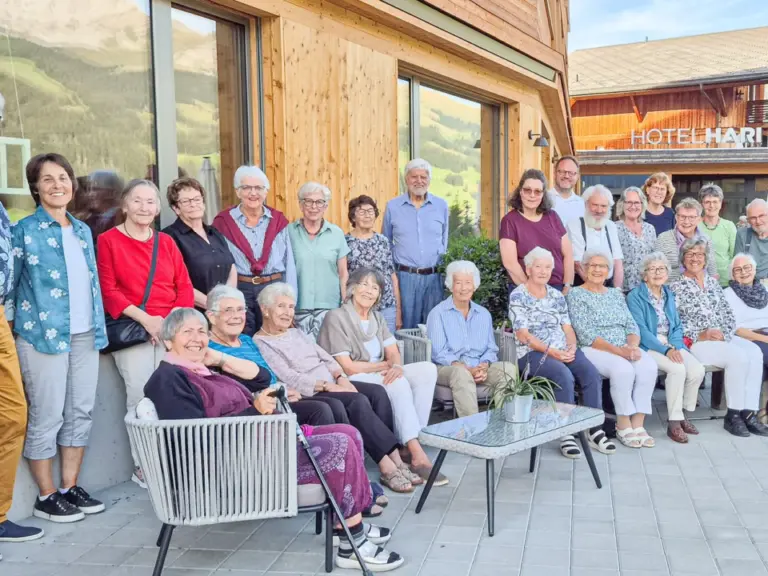 Grosse Gruppe älterer Menschen posiert auf einer Terrasse vor einem Berghotel in den Bergen.