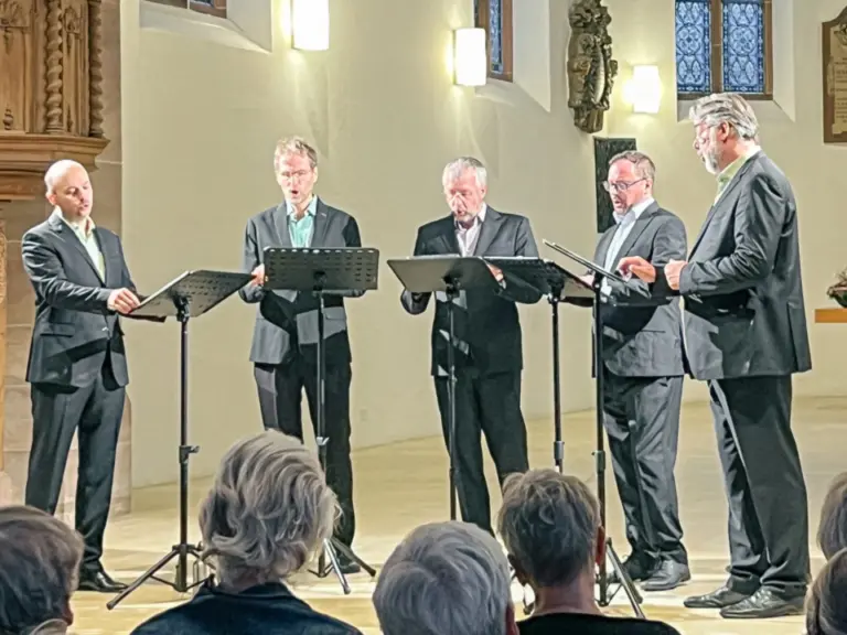Fünf Männer in Anzügen singen an Notenständern in einer Kirche vor sitzendem Publikum.