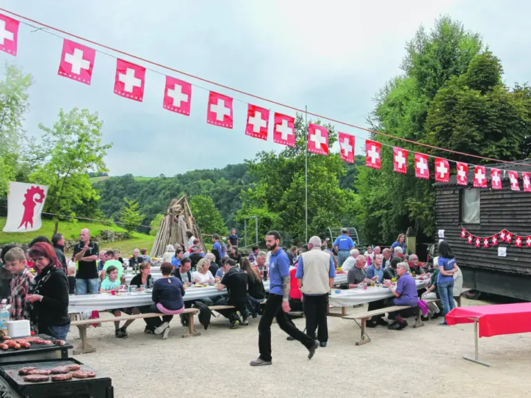 Menschen sitzen an Tischen unter Schweizer Fähnchengirlanden bei einem Fest im Freien, im Vordergrund grilliert jemand.