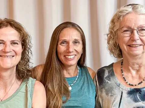 Drei lächelnde Frauen stehen nebeneinander vor einem hellen Vorhang, die mittlere hat die Arme um die anderen gelegt.