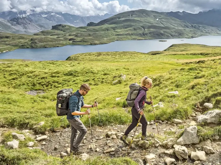 Zwei Wanderer mit Rucksäcken gehen über einen steinigen Pfad, im Hintergrund ein Bergsee und Gipfel.