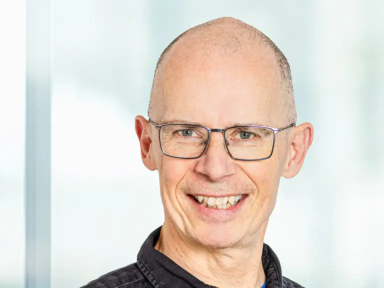 Ein lächelnder Mann mit Brille und kurzem Haar in dunklem Hemd blickt in die Kamera.