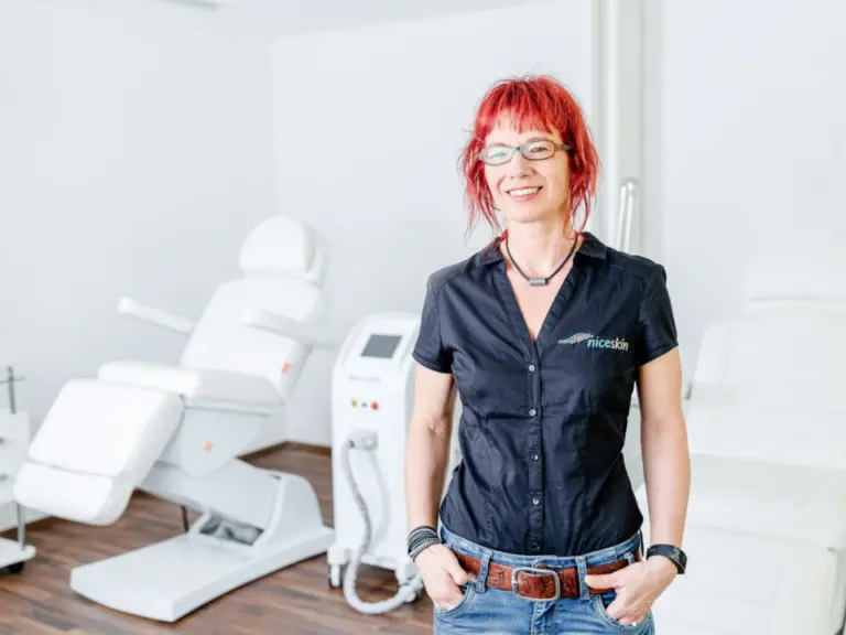 Rothaarige Frau mit Brille lächelt in einem hellen Behandlungsraum mit Kosmetikliegen.