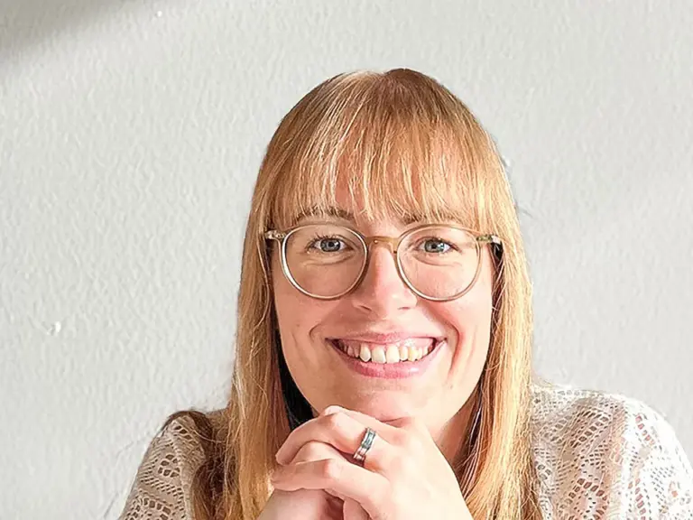 Eine lächelnde Frau mit runder Brille und roten Haaren faltet die Hände vor dem Kinn.