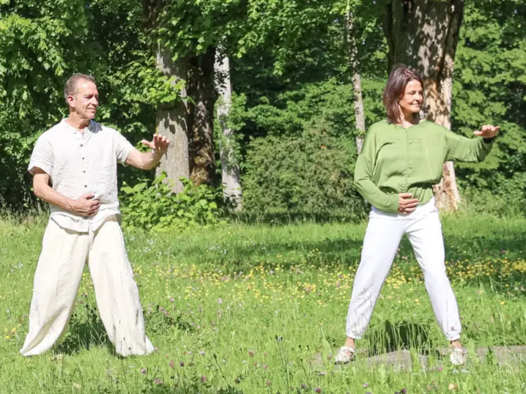 Ein Mann und eine Frau üben Qi Gong auf einer grünen Wiese zwischen Bäumen.