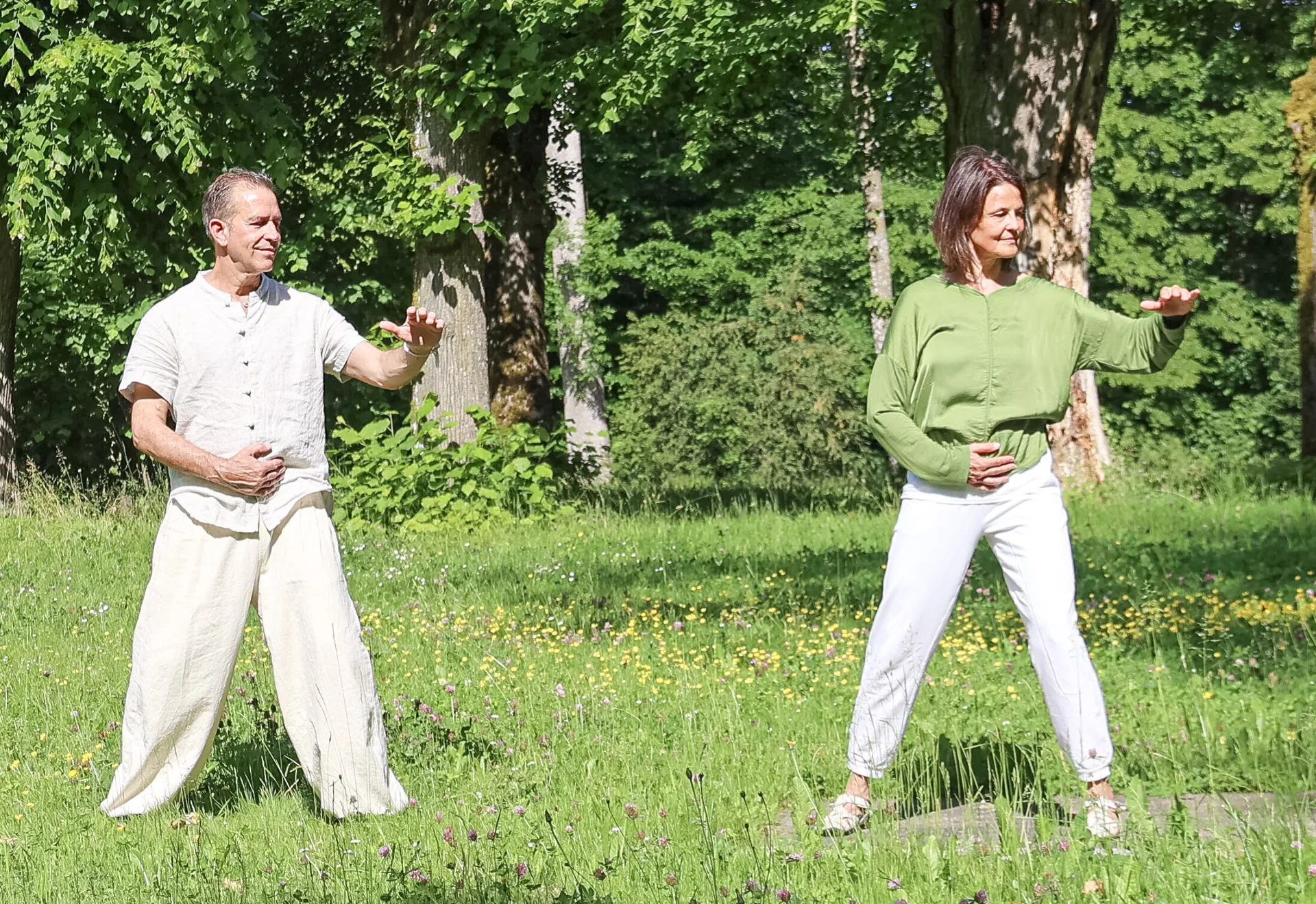 Ein Mann und eine Frau üben Qi Gong auf einer grünen Wiese zwischen Bäumen.