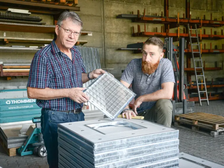 Zwei Männer in einer Werkstatt betrachten gemeinsam ein Metallgitter, im Hintergrund gestapelte Bleche.