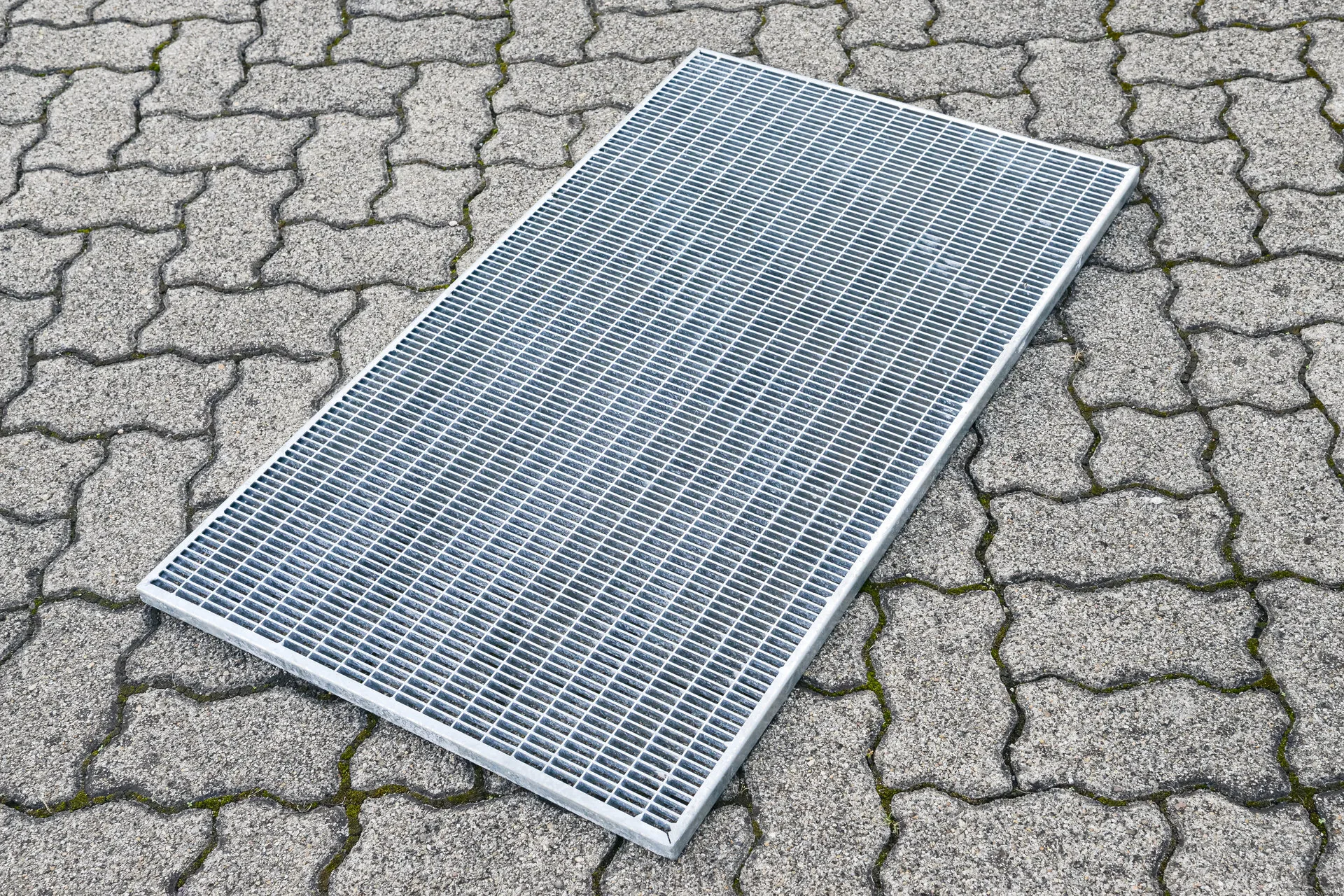 Ein rechteckiges Metallgitter liegt schräg auf einem gepflasterten Boden mit Moos in den Fugen.