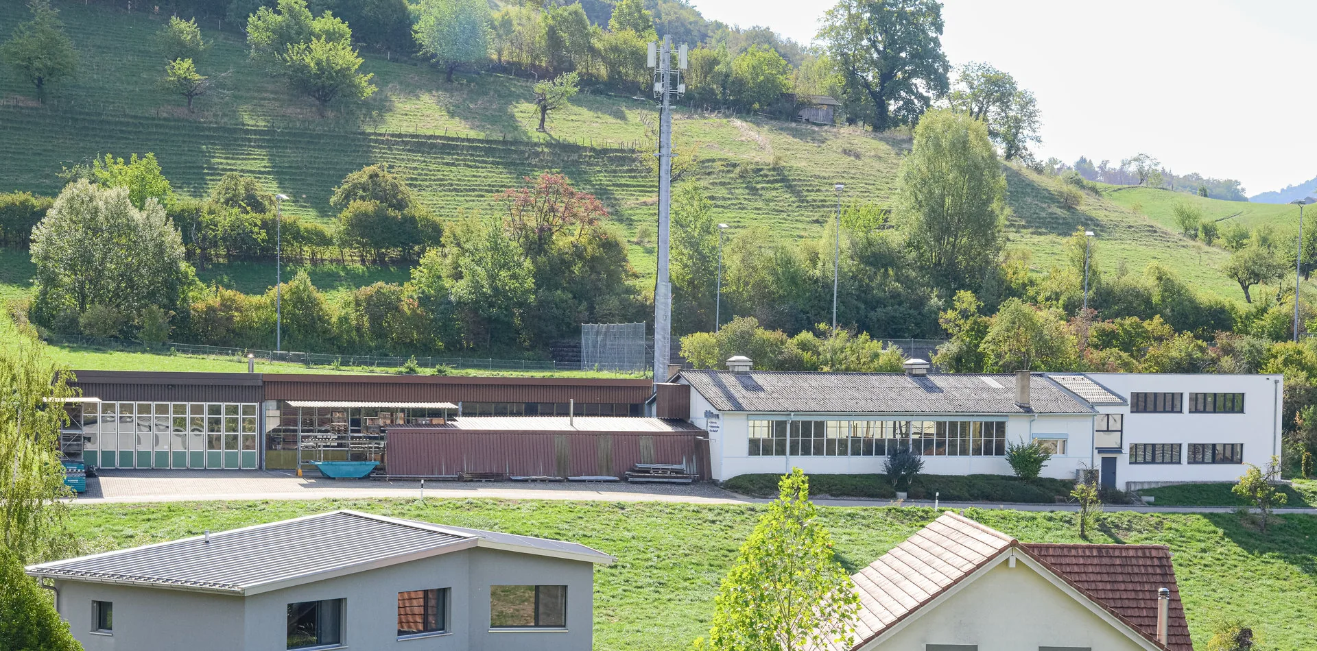 Zwei Wohnhäuser und ein langgezogenes Fabrikgebäude vor einem bewaldeten Hügel bei Sonnenschein.
