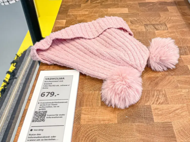 Ein rosa gestrickter Zipfelmütze mit Bommeln liegt auf einer Holzarbeitsplatte neben einem Preisschild.
