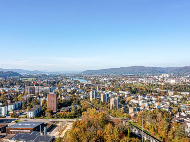Luftaufnahme einer Stadt mit Fluss, umgeben von bewaldeten Hügeln unter klarem Himmel.