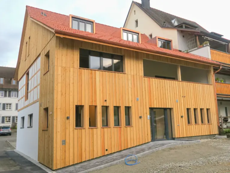Modernes Gebäude mit Holzfassade und rotem Ziegeldach neben einem älteren Fachwerkhaus.