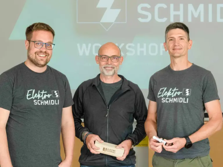 Drei lächelnde Männer stehen nebeneinander vor einer Präsentationsleinwand, der mittlere hält eine Auszeichnung.