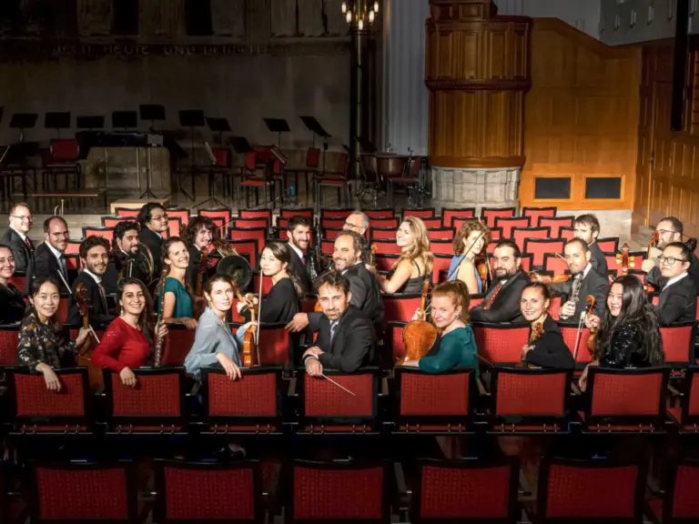 Ein Orchester mit Streichinstrumenten posiert in mehreren Reihen auf einer Bühne mit roten Stühlen.
