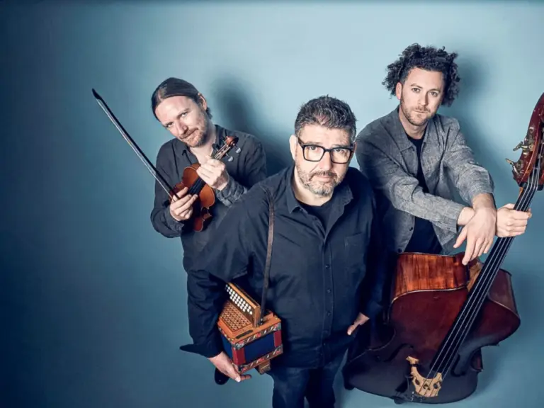 Drei Musiker vor blauem Hintergrund, mit Geige, Handorgel und Kontrabass posierend.