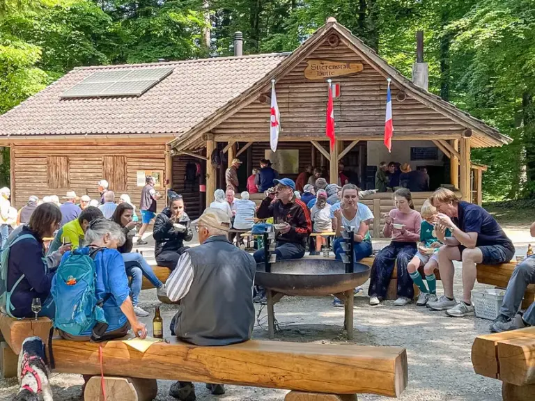 Menschengruppe sitzt auf Holzbänken vor einer Waldhütte im Wald und unterhält sich.