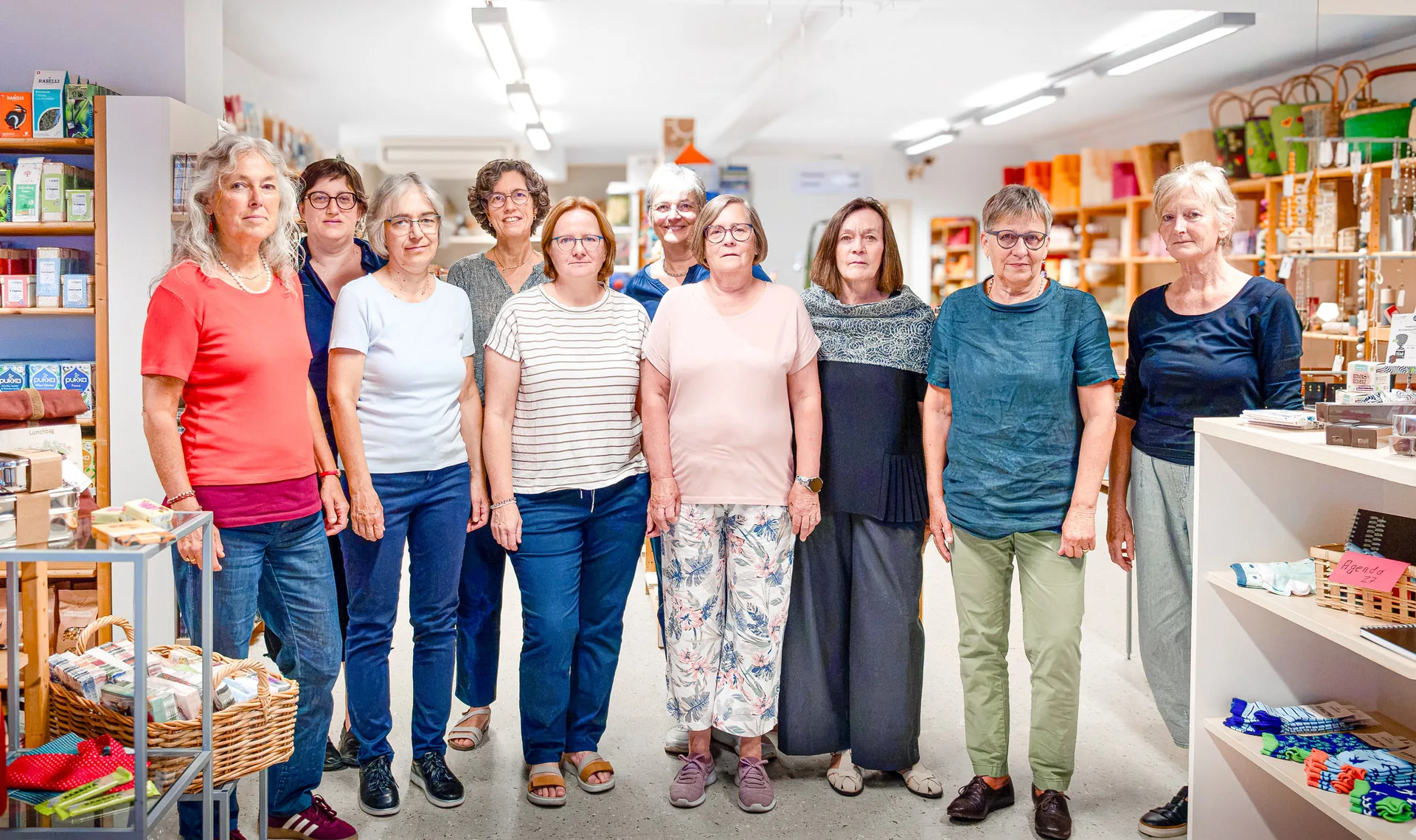 Eine Gruppe von neun Frauen steht nebeneinander in einem gut sortierten Laden mit Regalen voller Waren.