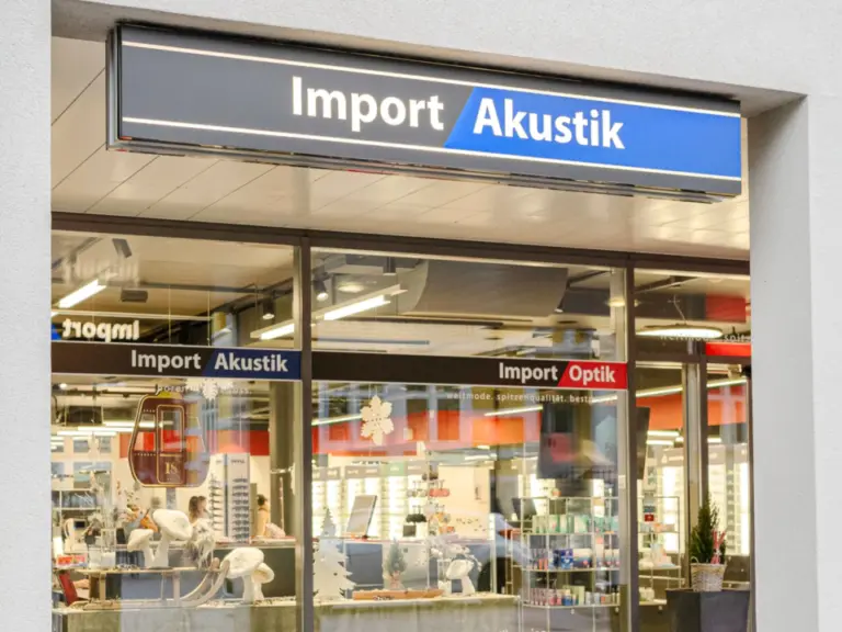 Ladenfassade mit blauem Leuchtschild „Import Akustik“ über grossen Schaufenstern eines Geschäfts.