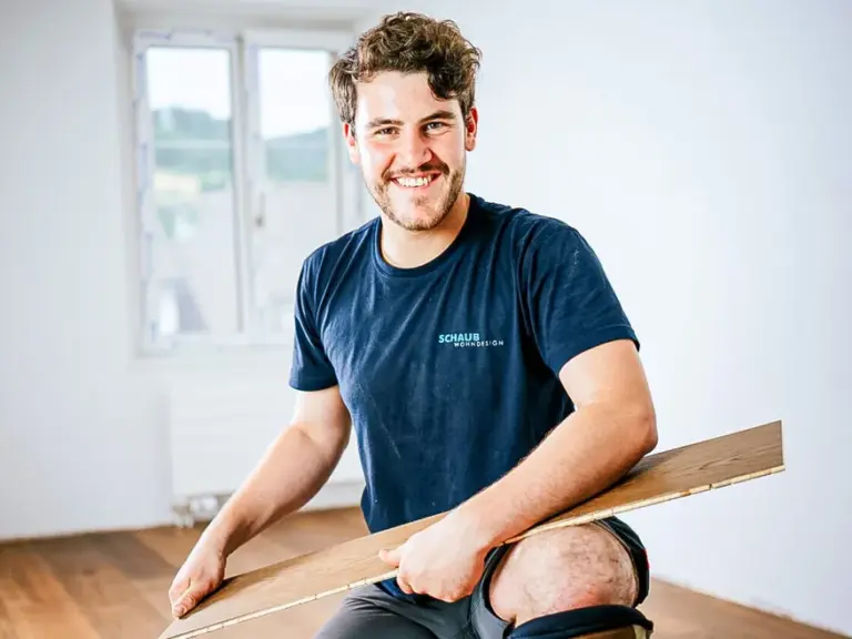 Ein lächelnder junger Handwerker kniet auf Knieschonern und hält ein Parkettbrett in den Händen.