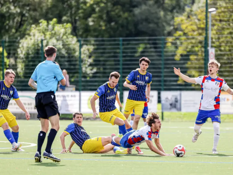 Fussballspieler in gelb-blauen und rot-blauen Trikots kämpfen um den Ball, ein Schiedsrichter beobachtet die Szene.