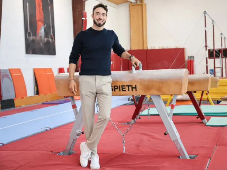 Ein Mann steht lächelnd in einer Turnhalle an einem Pauschenpferd, im Hintergrund Turngeräte und Matten.