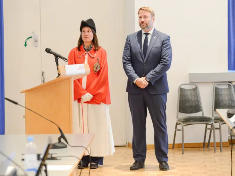 Ein Mann im Anzug steht neben einer Frau in traditioneller roter Amtstracht am Rednerpult im Saal.