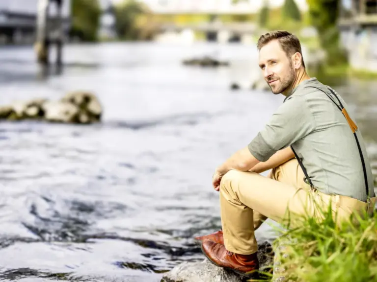 Ein Mann mit Hosenträgern sitzt am Flussufer auf einem Stein und blickt über das Wasser.