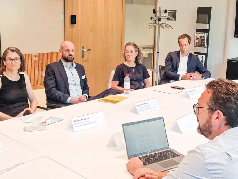 Mehrere Personen sitzen an einem ovalen Konferenztisch im Gespräch, im Hintergrund ein Bildschirm mit Porträts.
