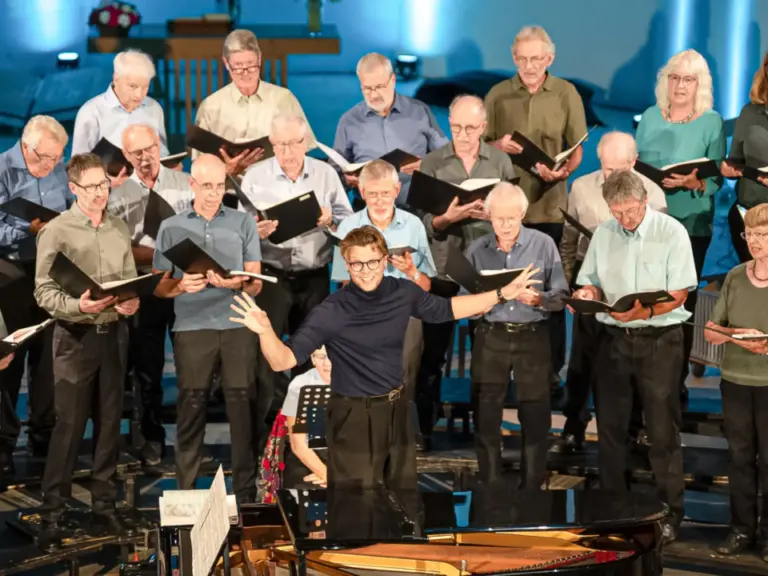 Ein Chor mit Noten in den Händen singt, während ein Dirigent mit ausgebreiteten Armen vor ihm steht.