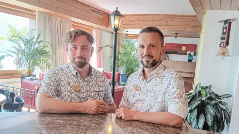 Zwei Männer in gemusterten Hemden stehen lächelnd hinter einer Steintheke in einem holzverkleideten Raum.