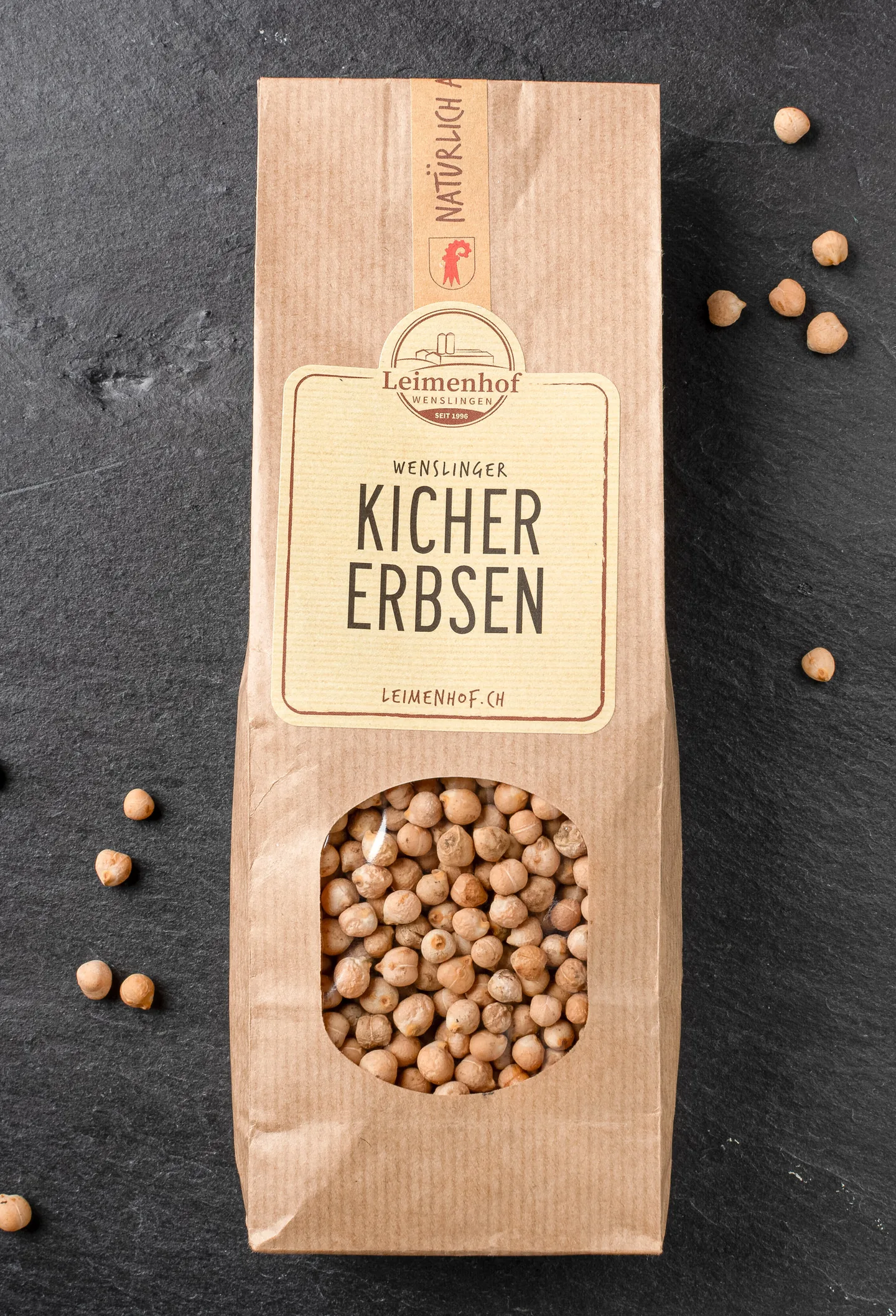 Eine Papiertüte mit Kichererbsen liegt auf dunklem Schieferuntergrund, einzelne Erbsen liegen verstreut daneben.