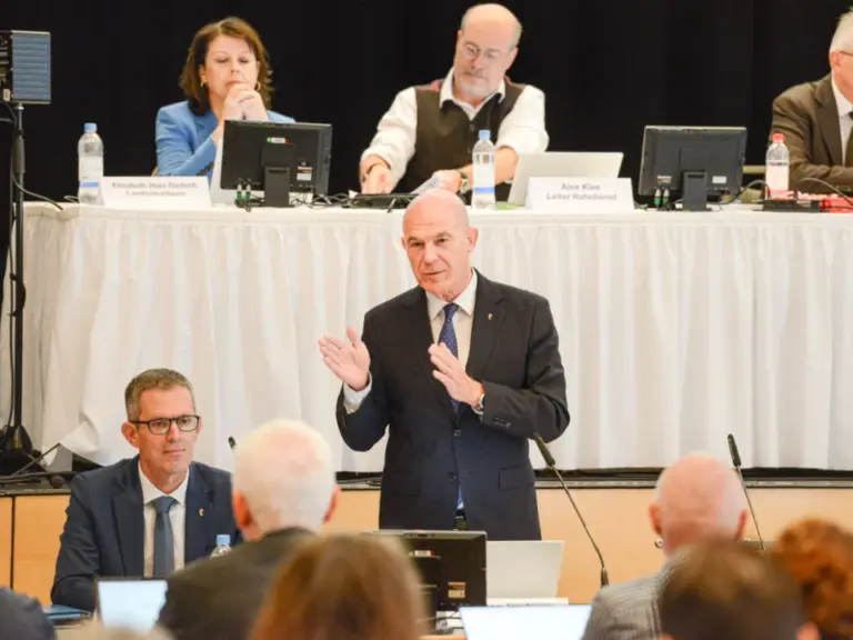 Ein Mann im Anzug steht redend mit erhobenen Händen am Mikrofon vor einem Saal.