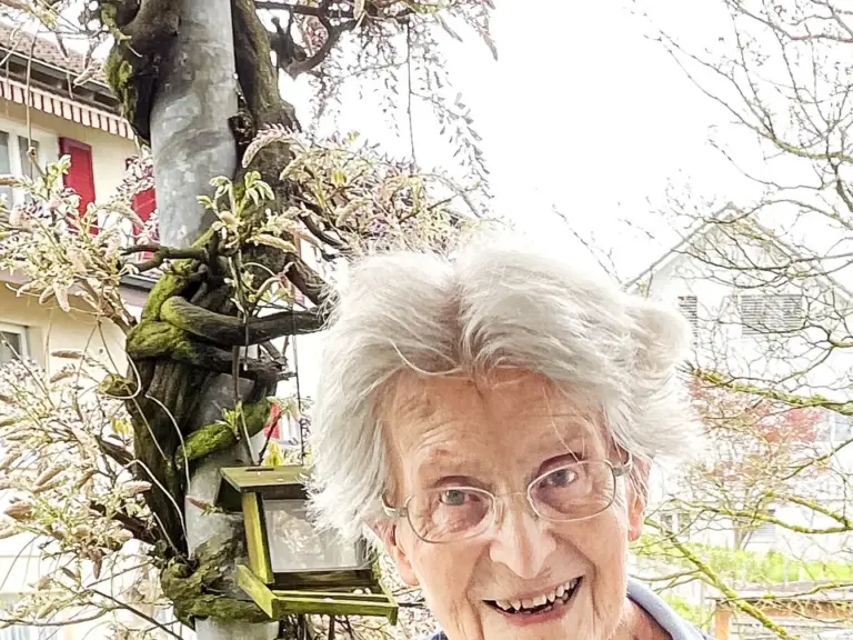 Ältere Frau mit weissen Haaren und Brille lächelt, sie trägt einen dunkelblauen Cardigan und stützt sich auf einen Rollator.